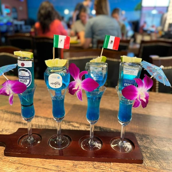 Four blue tequila shots with miniature bottles, limes, orchids, and Mexican flags on a wooden tray.