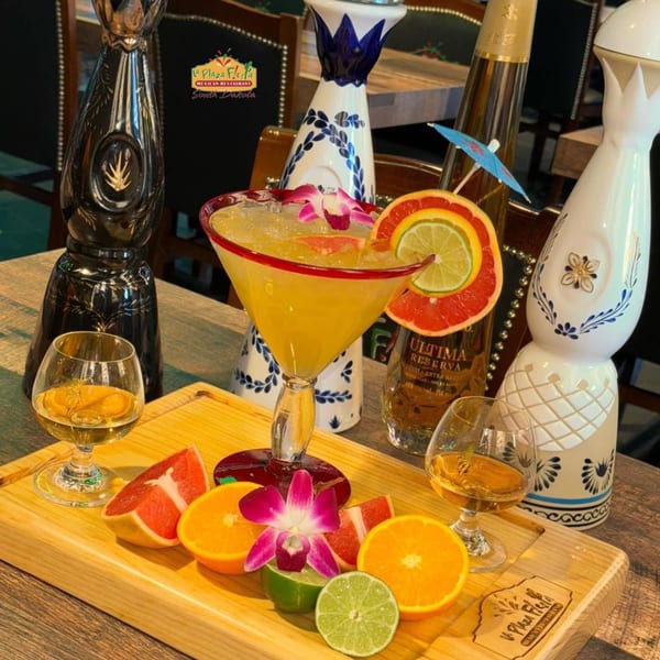 Margarita with fruit garnishes, tequila bottles, and glasses of amber liquid on a wooden board.