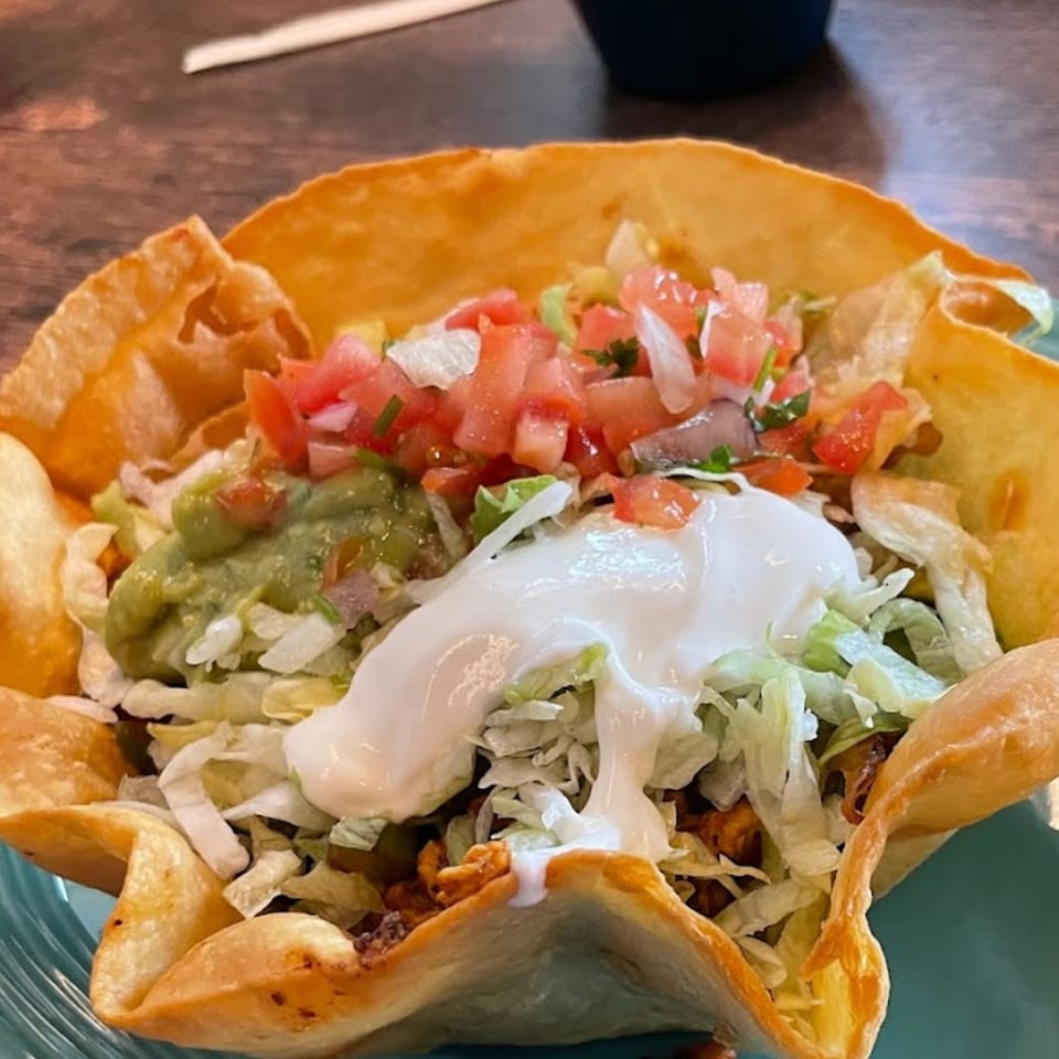 A taco salad in a crispy tortilla bowl, topped with guacamole, sour cream, lettuce, and pico de gallo.