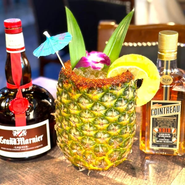 A pineapple cocktail with a lemon slice, orchid, and umbrella, flanked by Grand Marnier and Cointreau Noir bottles.