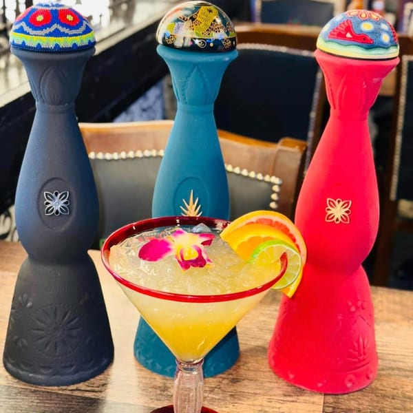 A vibrant La Plaza Fiesta margarita with fruit and orchid, flanked by three colorful tequila bottles.