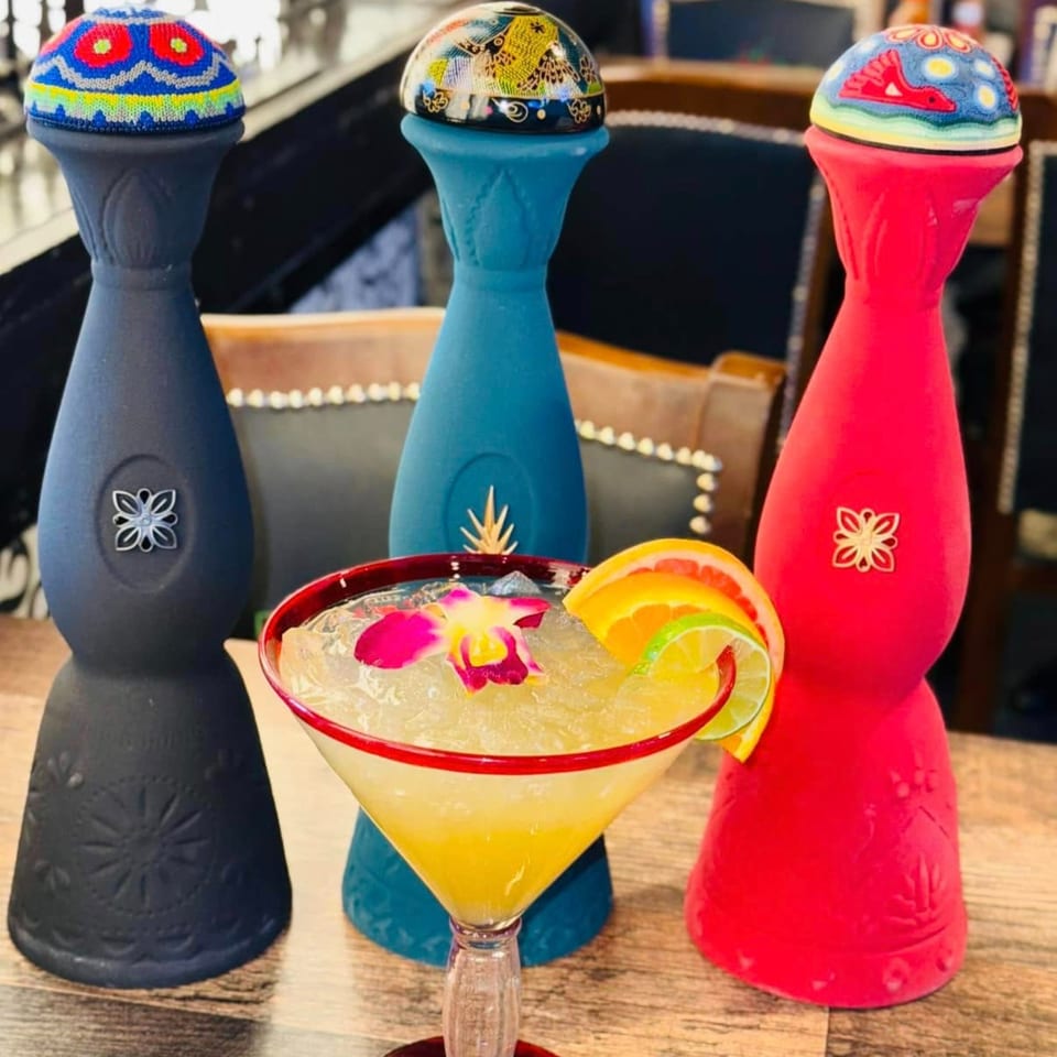 A vibrant La Plaza Fiesta margarita with fruit and orchid, flanked by three colorful tequila bottles.