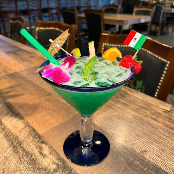 Green margarita with fruit, flower, umbrella, straw, and Mexican flag on a wooden bar.
