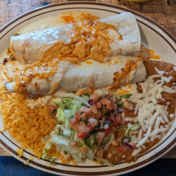 Two burritos covered in white and orange sauce, with rice, refried beans, and pico de gallo.