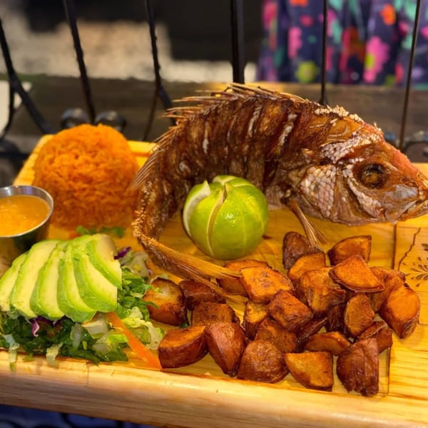 A whole fried fish with rice, avocado salad, fried plantains, and lime on a wooden board.