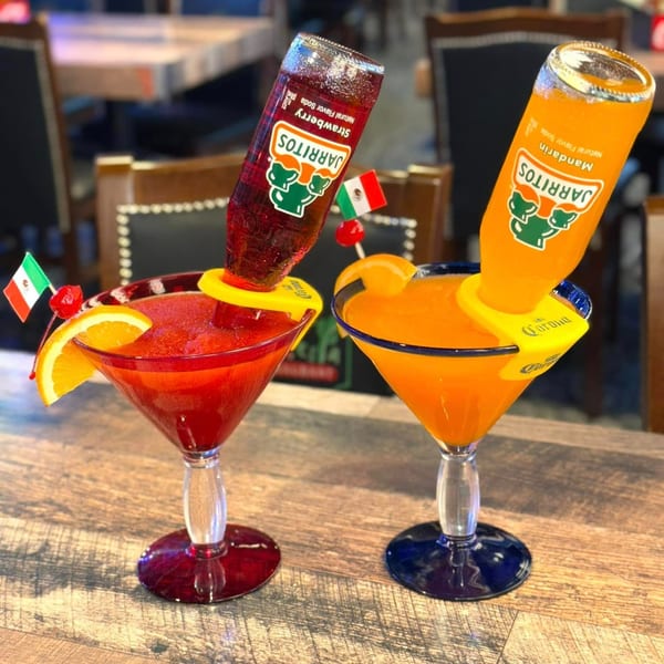 Two colorful Jarritos margaritas, strawberry and mandarin, garnished with fruit and Mexican flags.