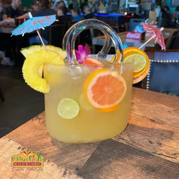 A large, clear handbag-shaped drink with fruit slices, ice, and two cocktail umbrellas on a wooden table.
